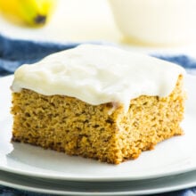 A slice of frosted vegan banana cake on a plate. In the background is more frosting and bananas.