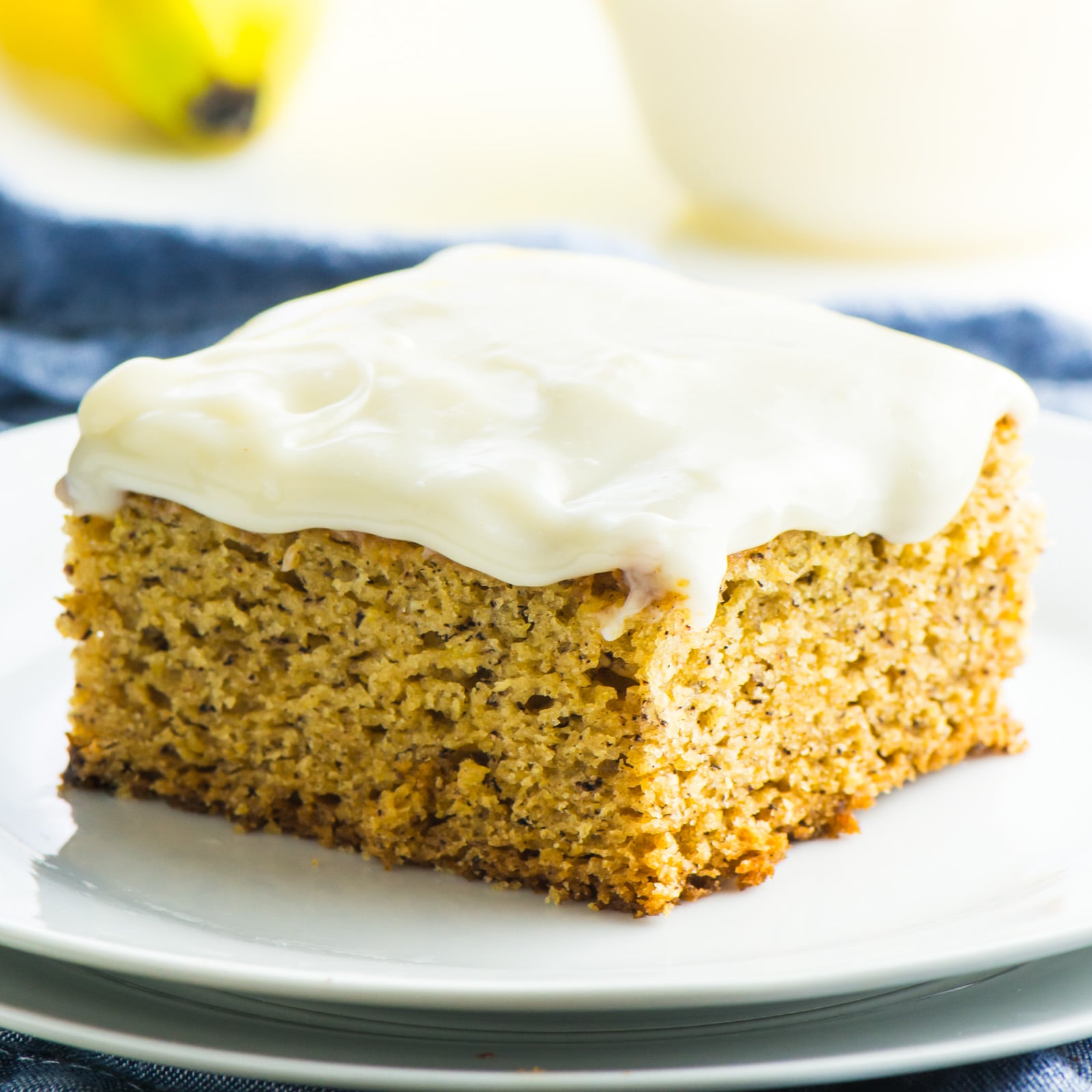 A slice of frosted vegan banana cake on a plate. In the background is more frosting and bananas.
