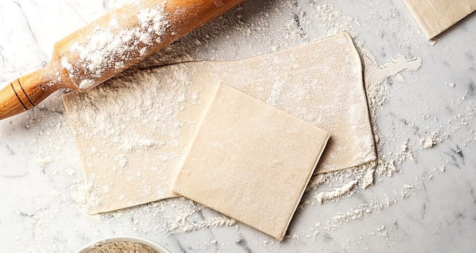 Sheets of puff pastry sit next to a floured wooden rolling pin on a marble counter top.