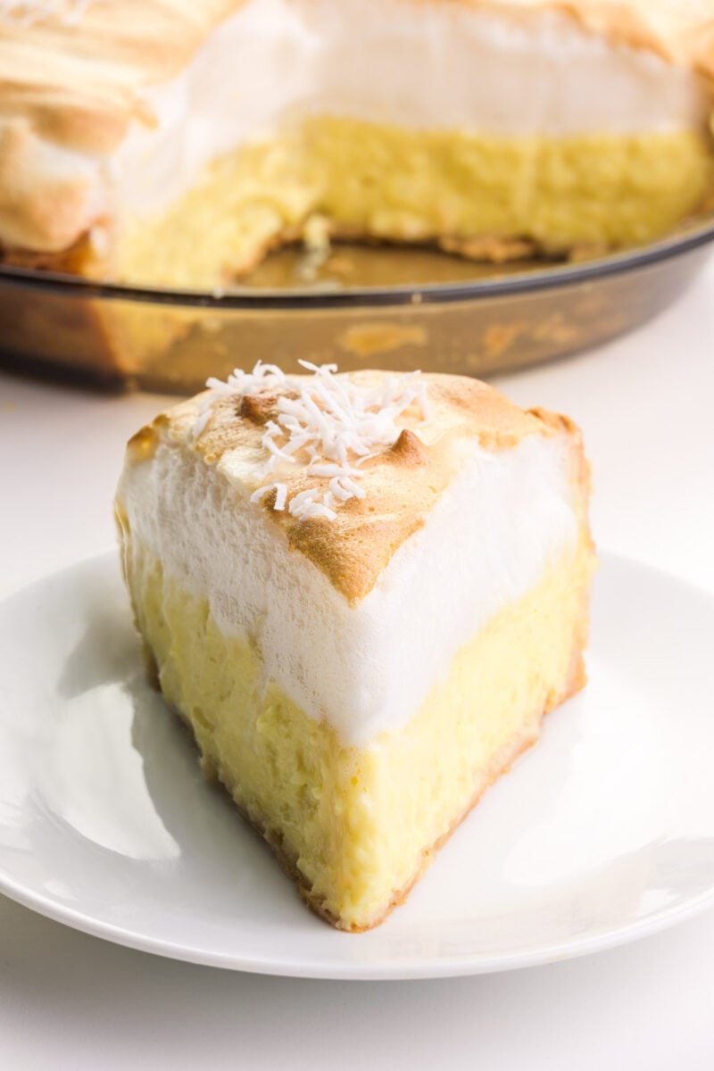 A slice of vegan coconut meringue pie sits o a plate. The rest of the pie is in the background.
