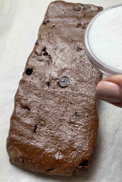 A hand holds sugar, sprinkling it over the top of an unbaked biscotti loaf.