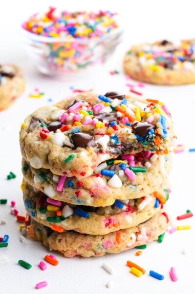 A stack of Vegan Chocolate Chip Sprinkle Cookies. The top cookie has a bite taken out and colorful sprinkles are scattered around.