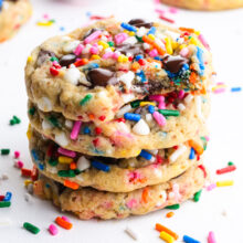 A stack of Vegan Chocolate Chip Sprinkle Cookies. The top cookie has a bite taken out and colorful sprinkles are scattered around.