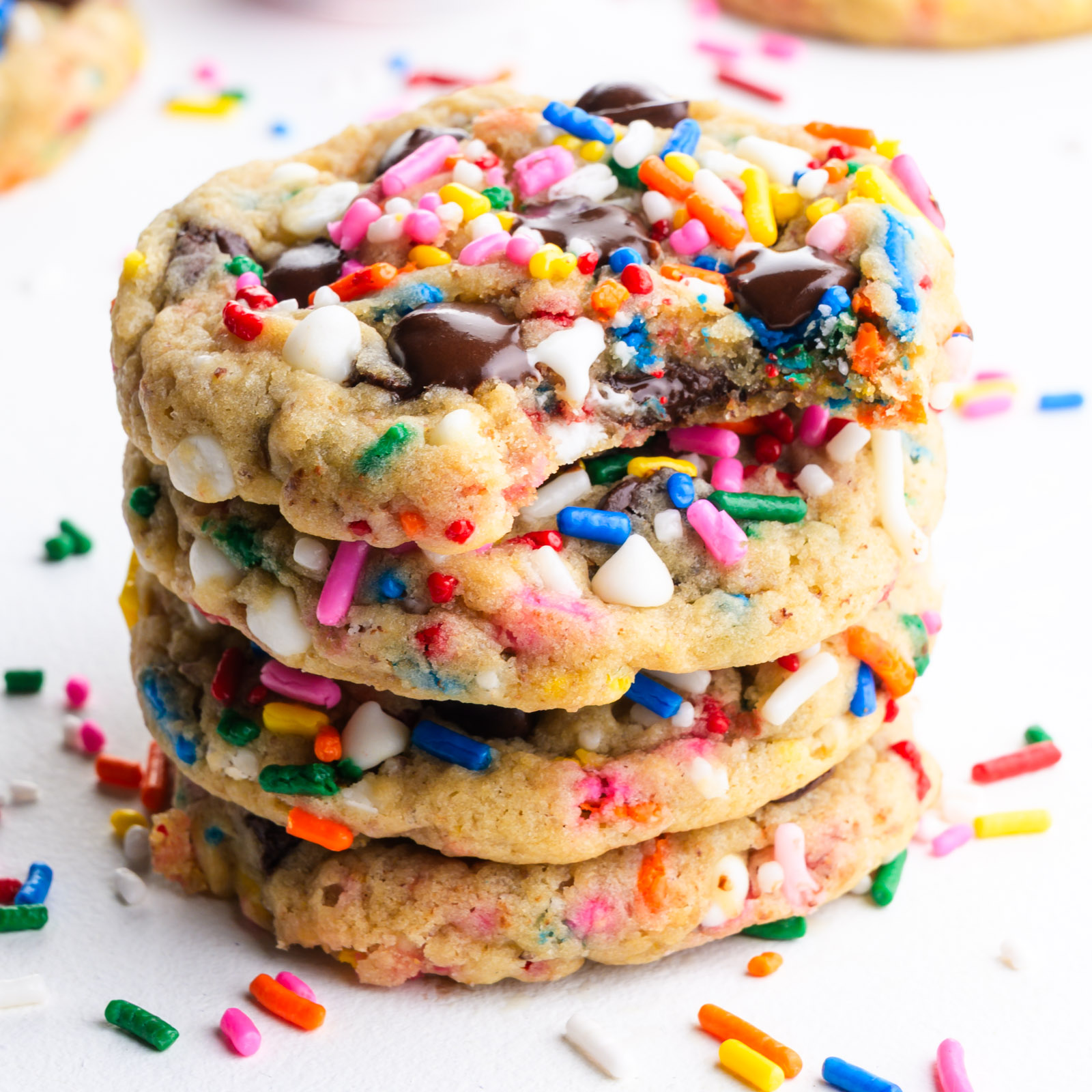 A stack of Vegan Chocolate Chip Sprinkle Cookies. The top cookie has a bite taken out and colorful sprinkles are scattered around.