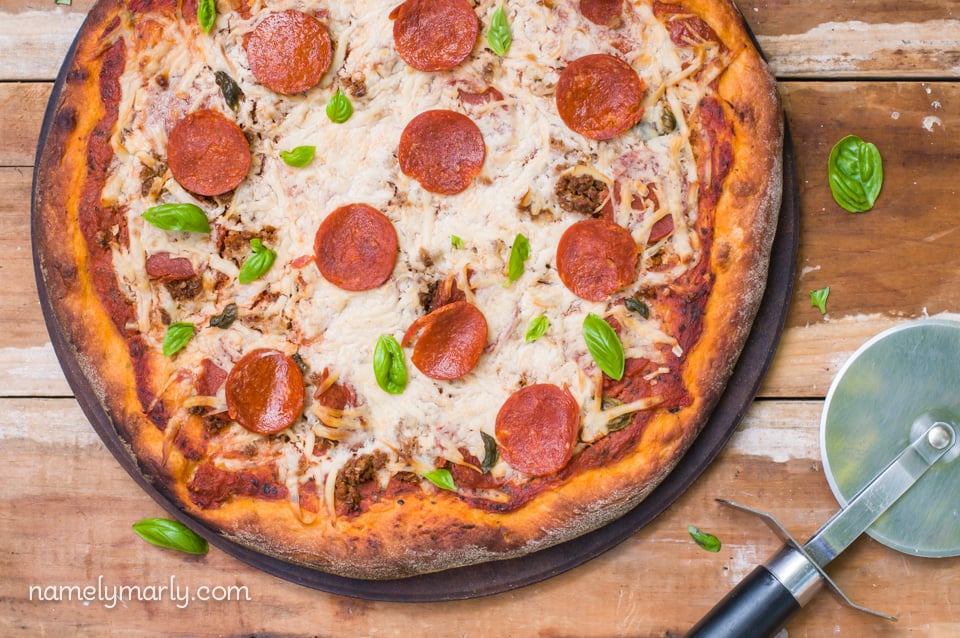 Looking down on a vegan pepperoni pizza. Fresh basil surrounds the pizza and a pizza cutters is shown to the right.