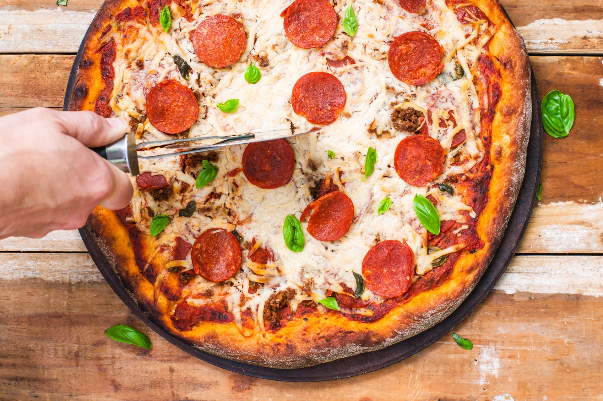 A hand reaches in with the pizza cutter to cut the pepperoni pizza.
