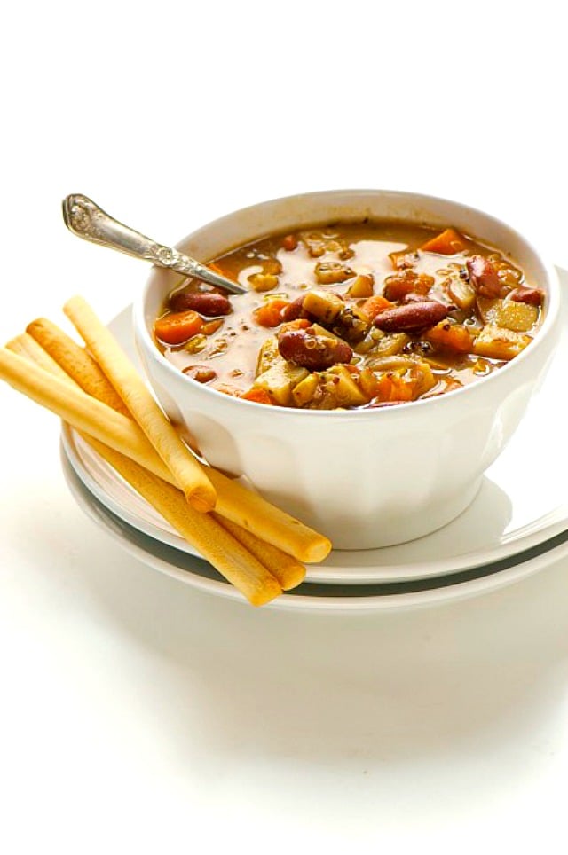 A bowl of gluten free minestrone with a spoon on it sits next to several breadsticks. 