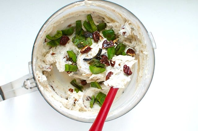 A creamy mixture in a food processor bowl. A red rubber spatula is being used to stir the ingredients. There are nuts and herbs added to the mixture as well.