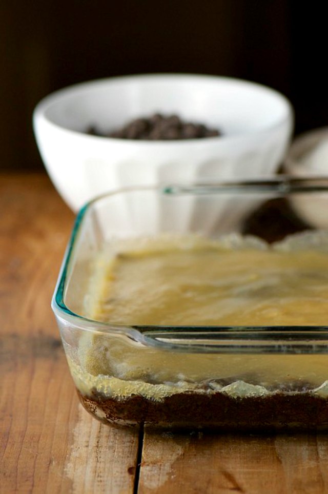 Vegan condensed milk is spread over vegan brownies in a baking dish. A bowl of chocolate chips is behind it.