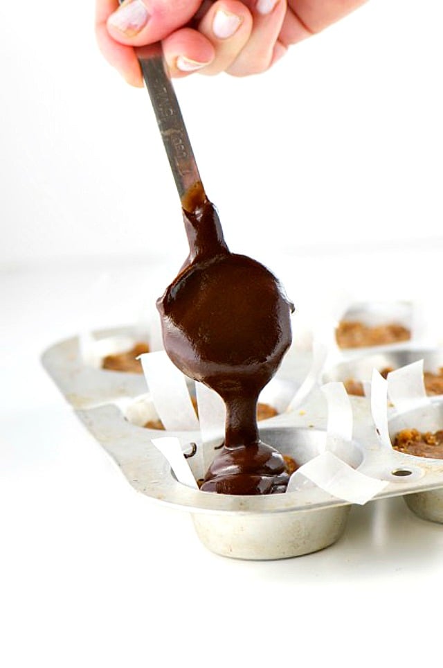 A hand holds a spoon drizzling melted chocolate over peanut butter in mini muffin molds.
