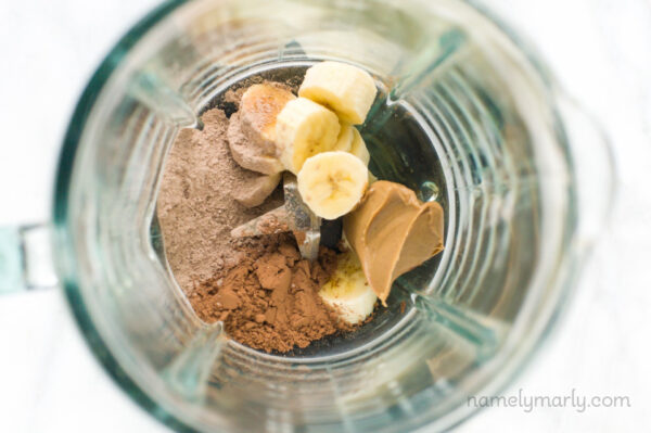 Looking into a blender jar there are ingredients like banana slices, chocolate protein powder, peanut butter, and more.