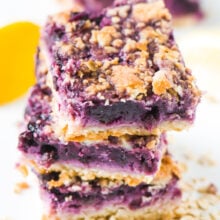 A stack of three vegan blueberry lemon pie bars are surrounded by oatmeal, lemons, and more slices in the background.