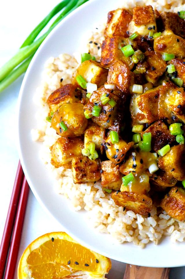 Crispy Orange Tofu sits in a white plate with chopsticks nearby and green onion at the top.