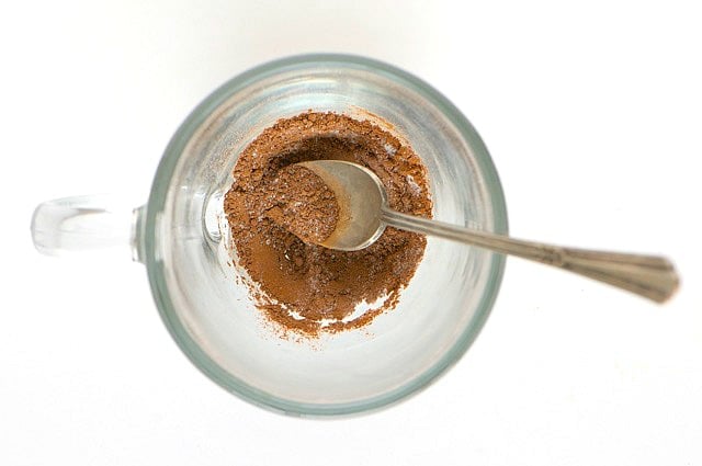 Looking down on the cocoa mix at the bottom of the glass coffee mug. A spoon is in the cup as well.