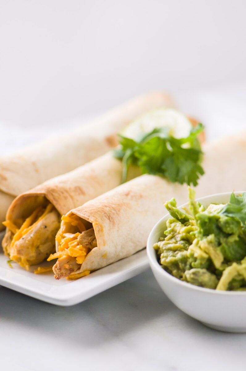 A plate holds vegan taquitos with herbs on top. Theres a bowl of mashed avocados on the side.