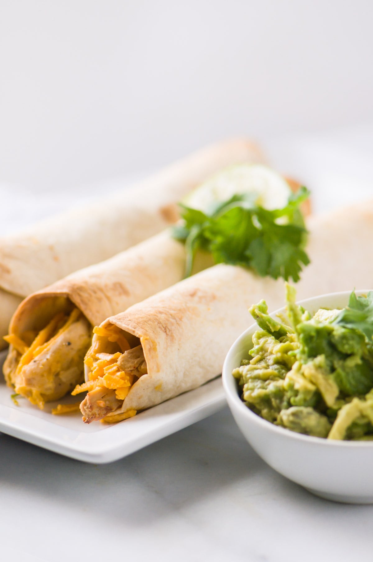 A plate holds vegan taquitos with herbs on top. Theres a bowl of mashed avocados on the side.