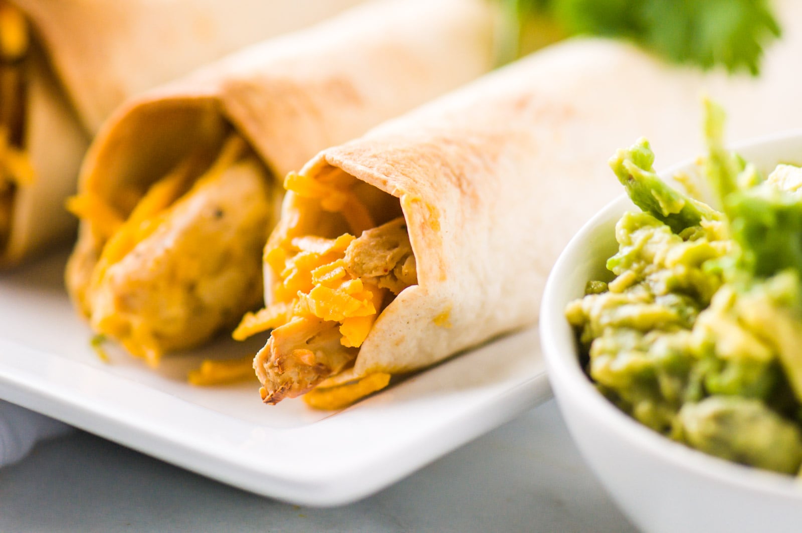 A closeup shot of rolled tacos with melted cheese sit on a plate. There is a bowl of mashed avocados beside it.