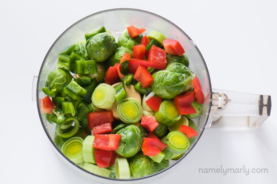 Looking down on a food processor full of raw Brussels sprouts, red bell peppers, and sliced jalapeños. 