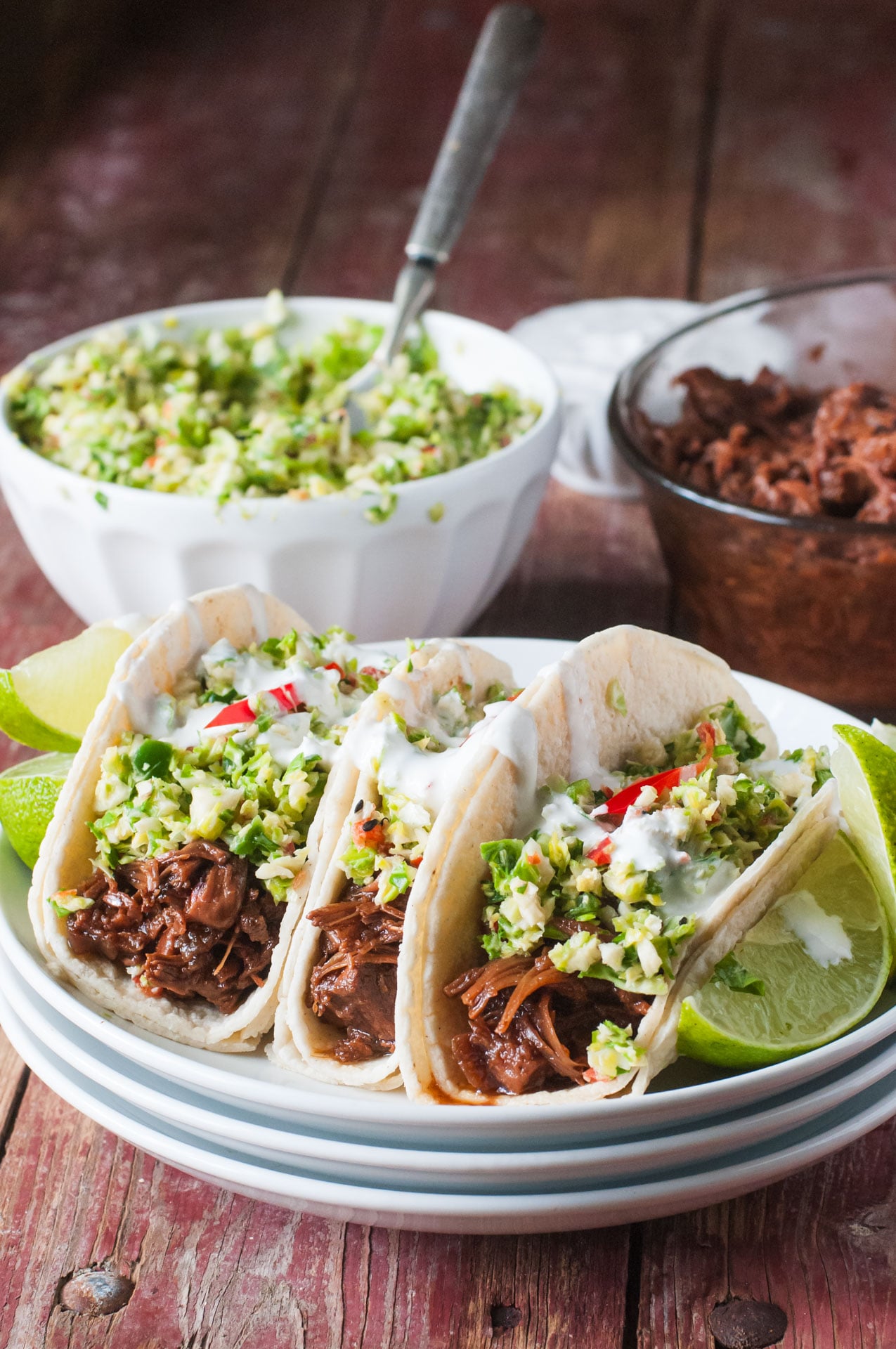 Three Bulgogi Jackfruit Street Tacos on a plate, topped with slaw. There's lime wedges on both sides, slaw and more bulgogi jackfruit behind it.