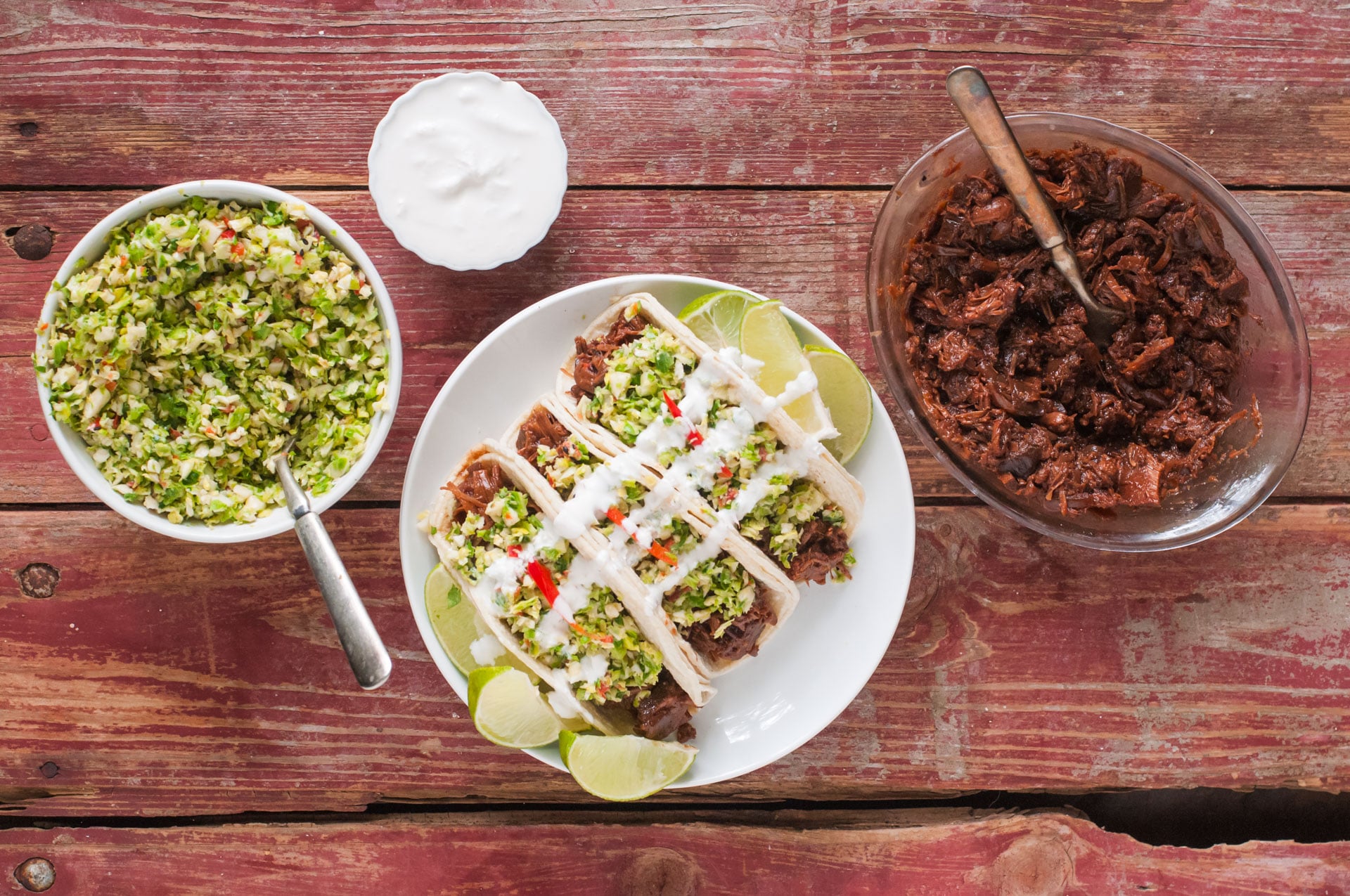 Looking down on three Bulgogi Jackfruit Street Tacos on a plate. Beside it are bowls of slaw, sour cream sauce, and more bulgogi jackfruit.