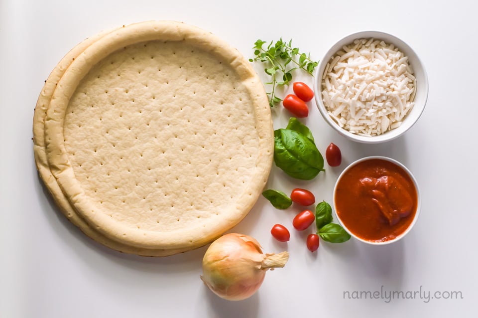 Looking down on two pizza crusts with bowls of vegan mozzarella shreds, pizza sauce, an onion, fresh herbs, and cherry tomatoes.