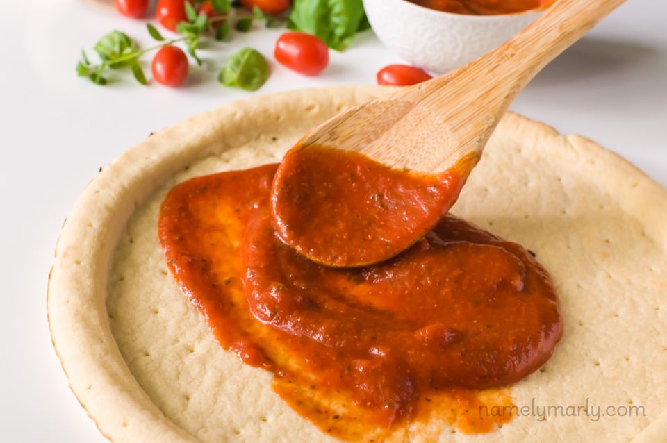 A wooden spoon is spreading pizza sauce over a crust. There is more sauce, basil leaves, and cherry tomatoes in the background.