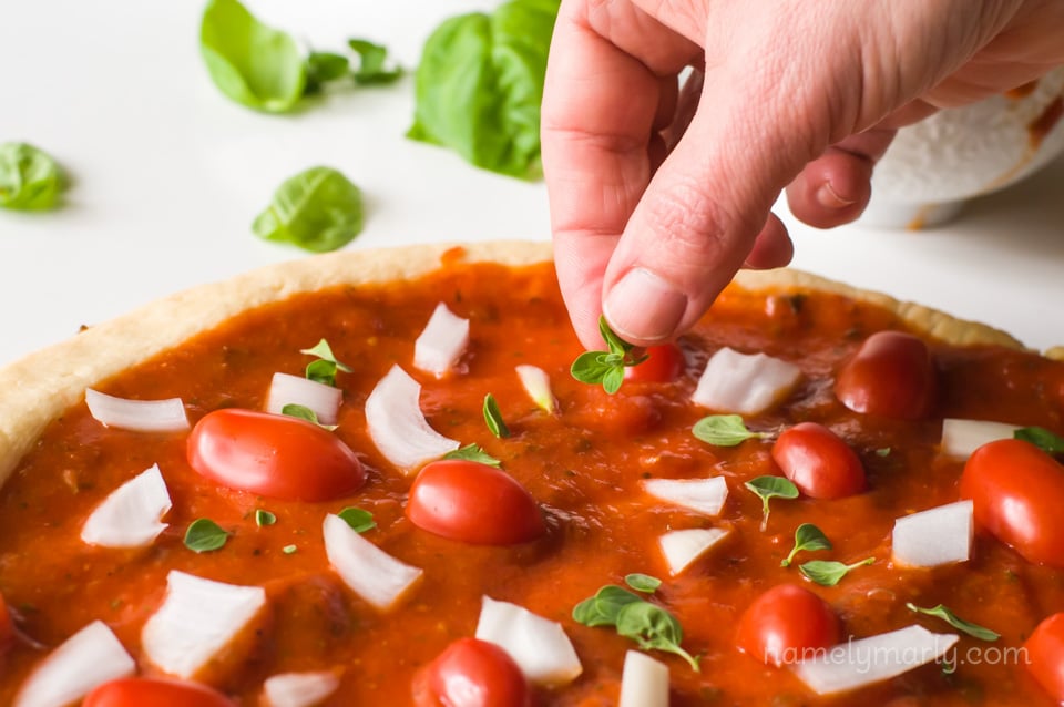A hand is placing fresh herbs on a pizza crust. There are more herbs in the background.