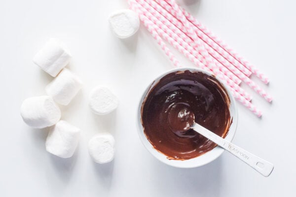 A bowl of melted chocolate sits next to vegan marshmallows and pink paper straws.