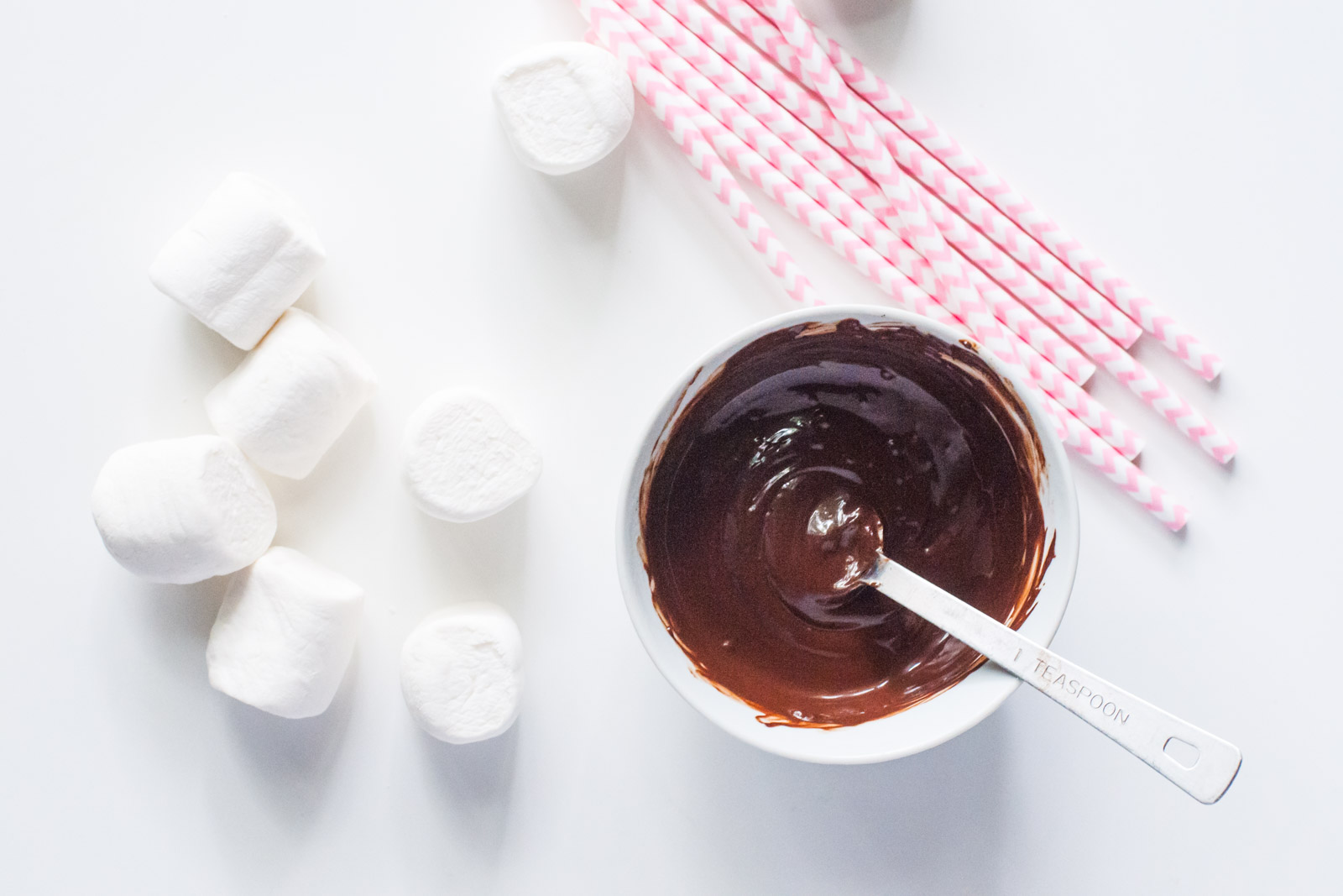 A bowl of melted chocolate sits next to vegan marshmallows and pink paper straws.