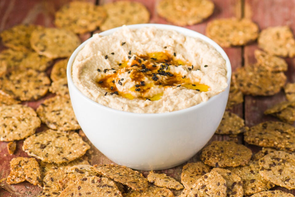 A bowl of creamy dip is topped with smoke sauce, black sesame seeds, and olive oil. It's surrounded by crackers.