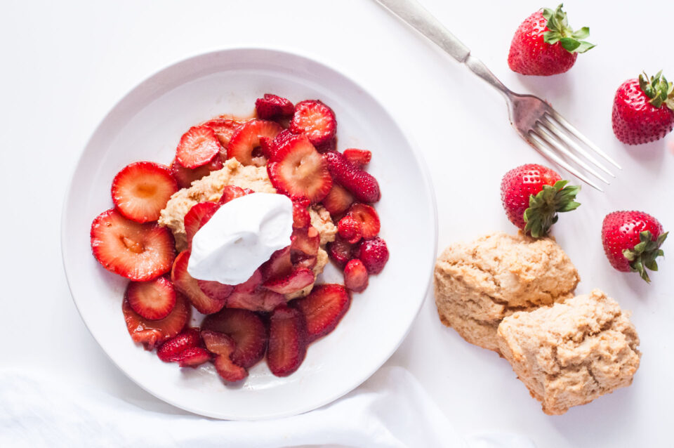 Looking down on vegan strawberry shortcake piled high with strawberries and whipped cream. More biscuits and berries are beside it.