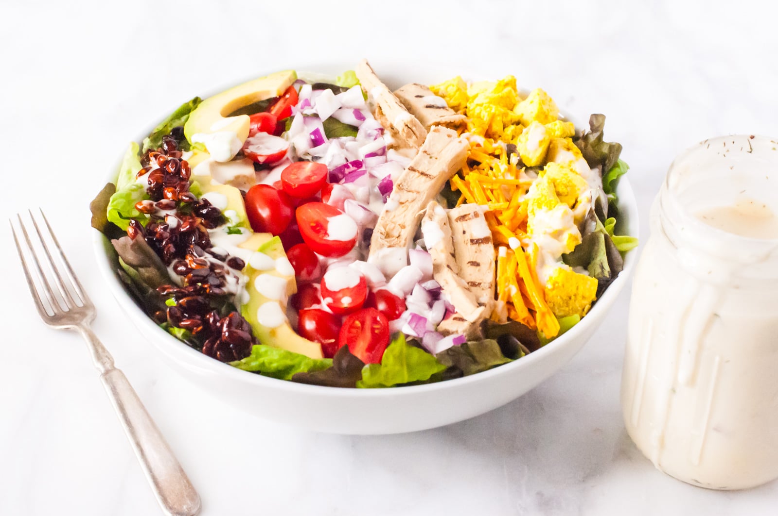 A vegan salad is in a white bowl. There's a creamy dressing in a jar next to it and a fork on the other side.