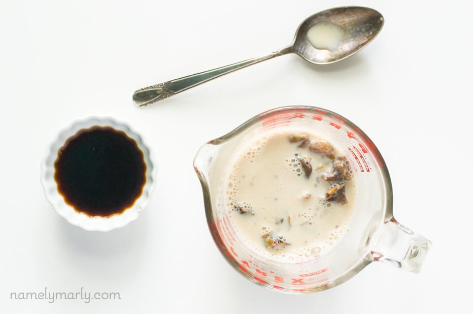 A pyrex measuring cup holds vegan milk with dates. Next to that is a small white bowl with liqueur. A spoon sits above this.