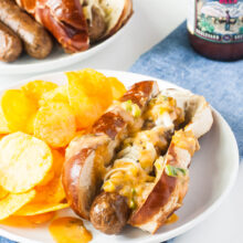 A vegan reuben brats is on a plate with potato chips beside it. A bottle of beer is behind it along with a plate full of more brats.