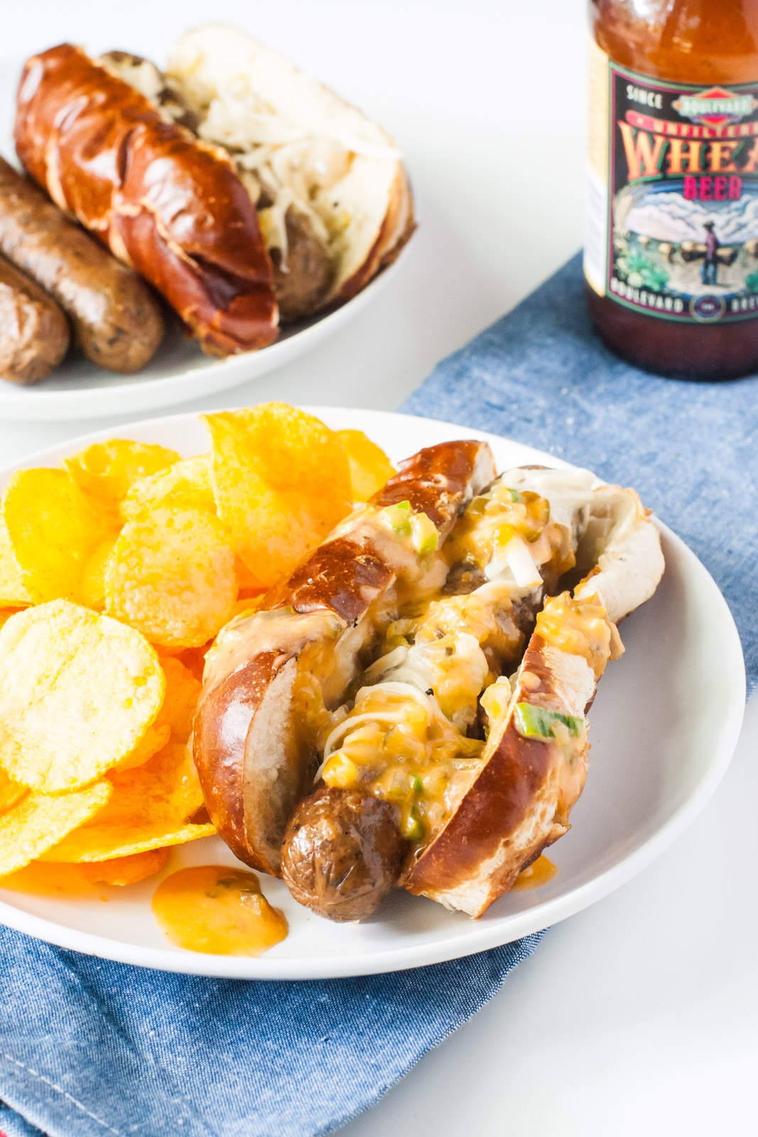 A vegan reuben brats is on a plate with potato chips beside it. A bottle of beer is behind it along with a plate full of more brats.