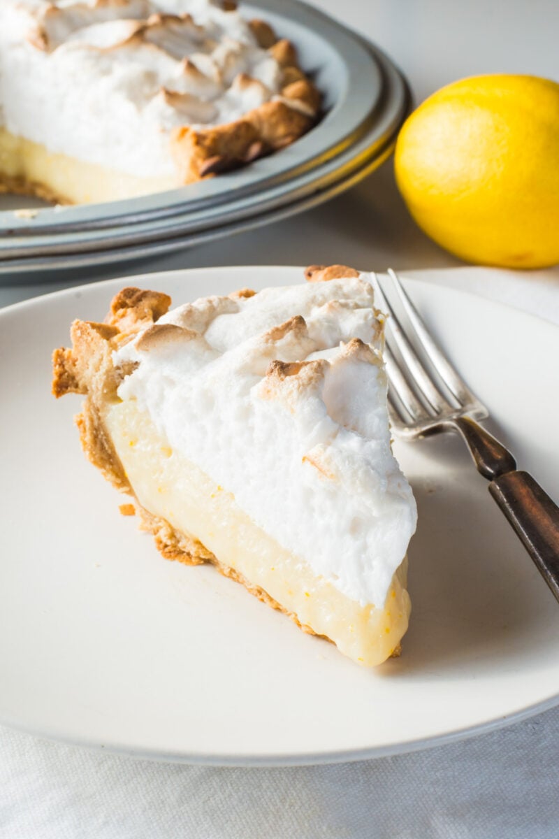 A slice of lemon pie on a plate with a fork beside it. A lemon and the rest of the pie is behind it.
