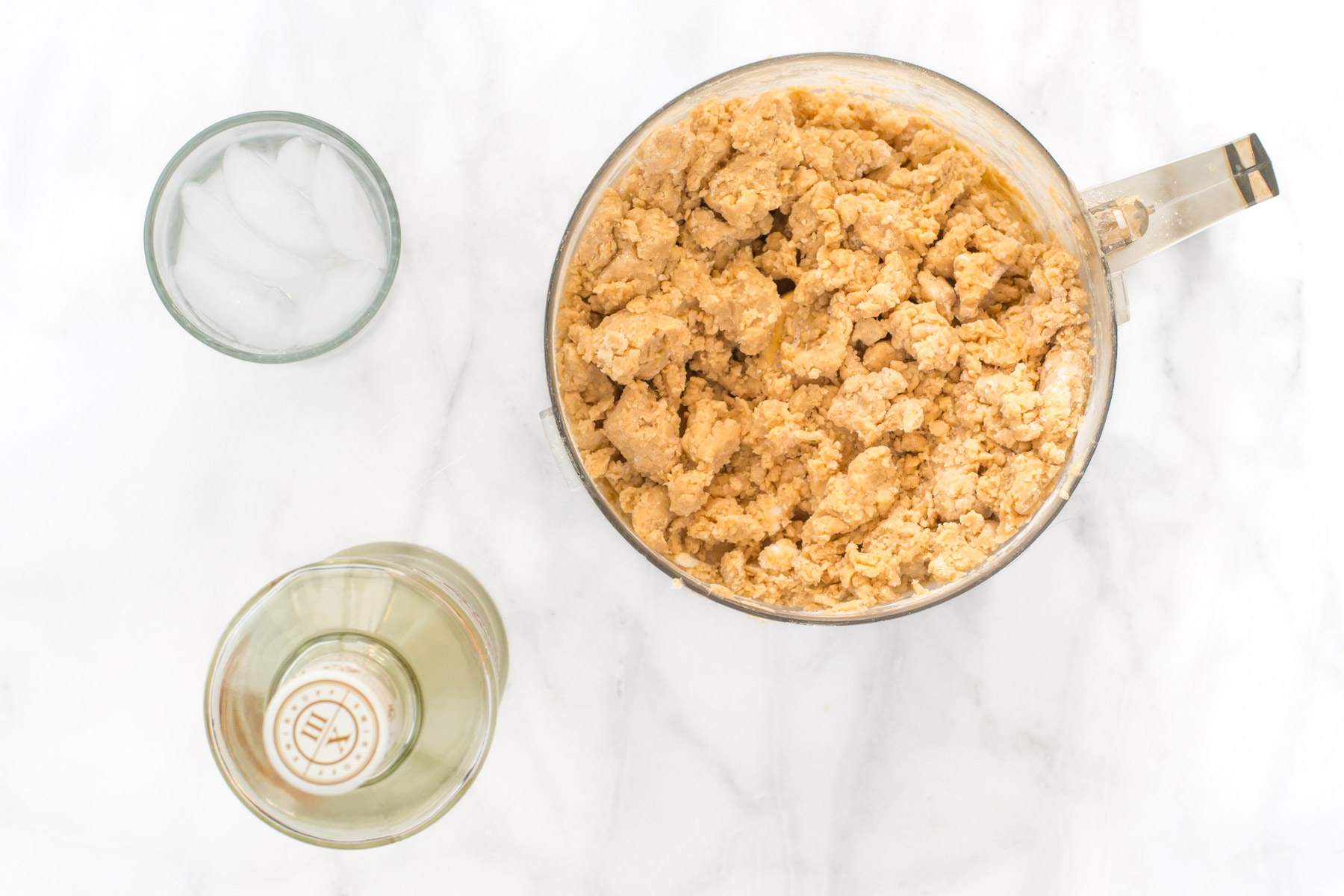 A food processor is full of crumbly pie crust mixture. A glass of ice water and bottle of vodka sit next to it.