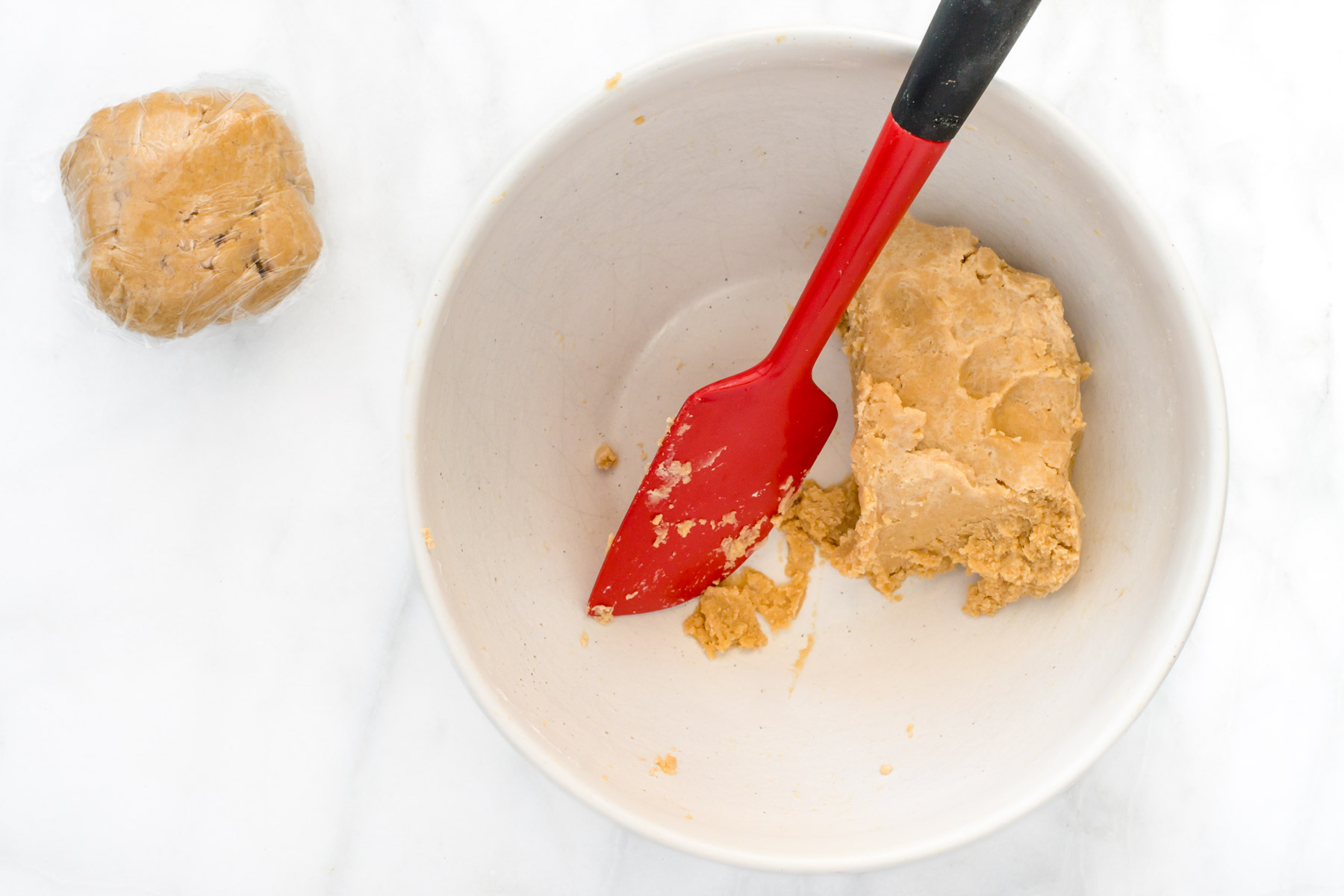 A bowl shows some dough mixture with a red spatula. A ball of dough wrapped in plastic sits beside the bowl.
