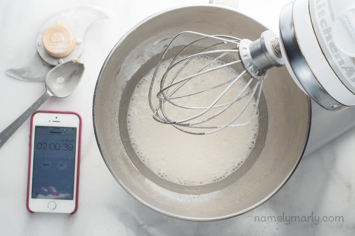 Looking down on a stand mixer and the mixture in the bottom is turning white. A phone next to the mixer indicate it's been beating for 2 minutes.