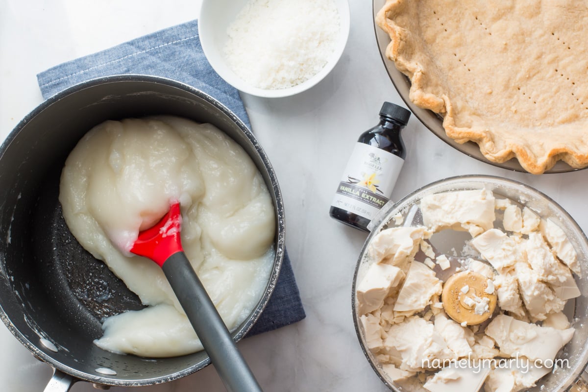 Looking down on the ingredients for making a pie, including tofu in a food processor, a baked pie crust, and the pudding in a saucepan. 