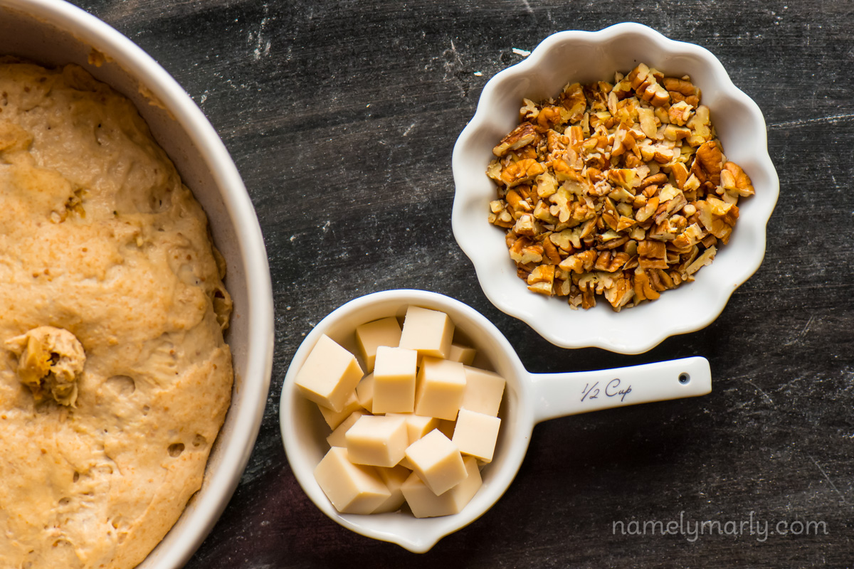 Looking down on a bowl of bread dough rising, a bowl of vegan cheese cubes, and chopped pecans.