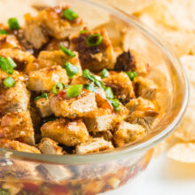 A serving dish full of vegan rangoon dip is topped with sliced green onions. There are tortilla chips beside it.