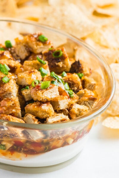A serving dish full of vegan rangoon dip is topped with sliced green onions. There are tortilla chips beside it.