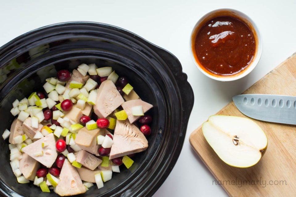 The ingredients in a slow cooker to make BBQ jackfruit. There's a bowl of BBQ sauce and a sliced pear beside it.