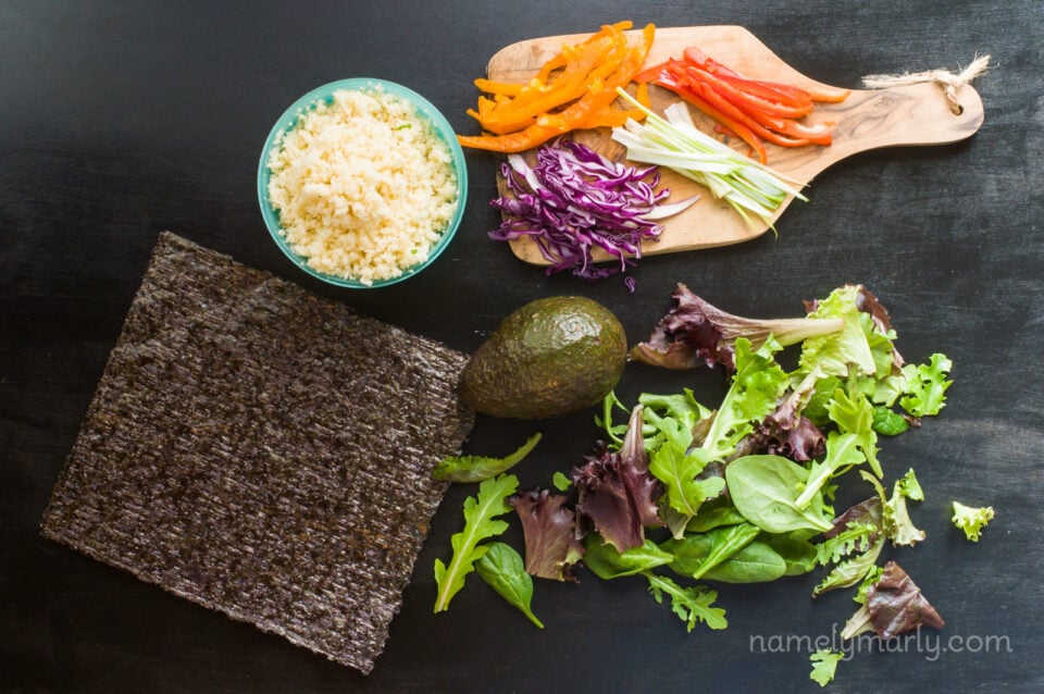 Sheets of nori sit next to an avocado, cauliflower rice, field greens, and a cutting board with sliced veggies.