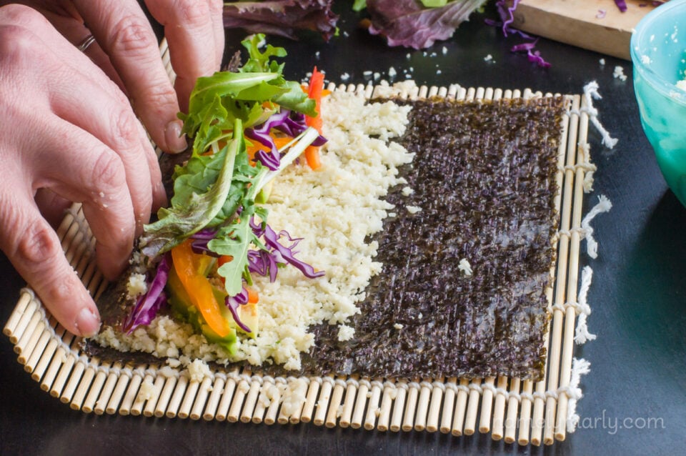Hands are beginning to roll a sheet of nori with veggies on top on a bamboo matt.