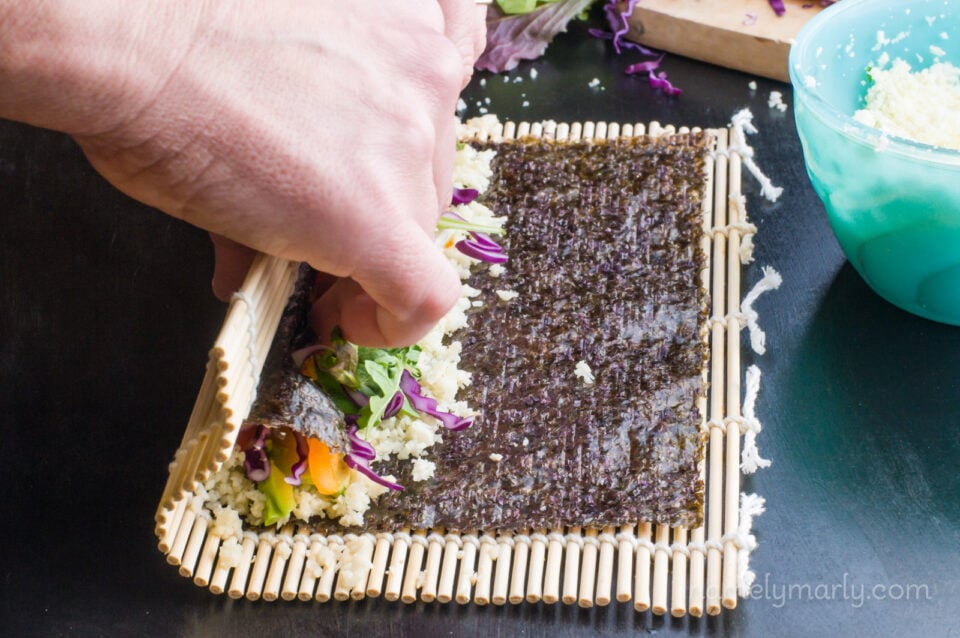 Hands continue to roll the nori topped with sliced veggies, sitting on a bamboo mat.