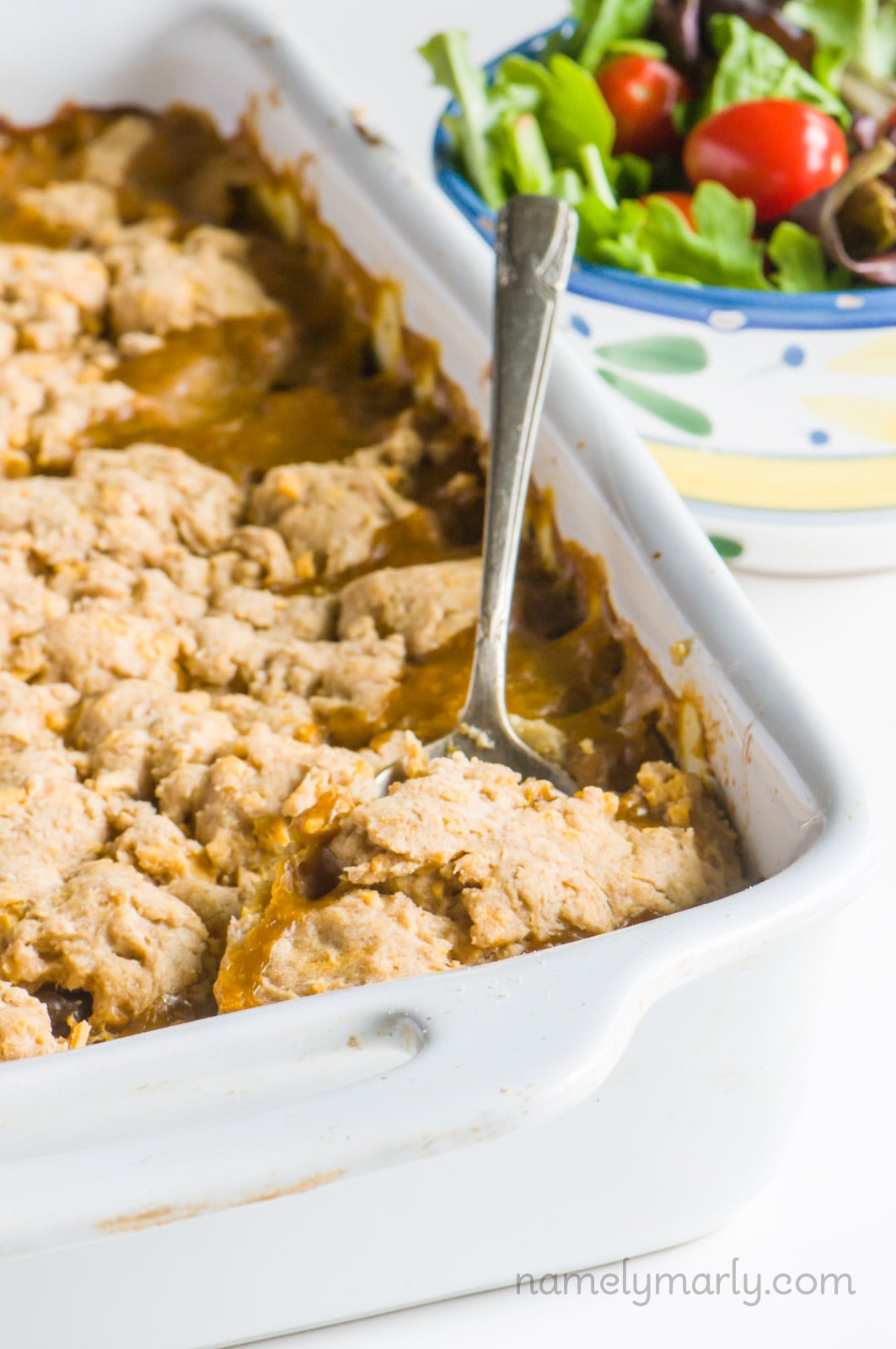 A casserole dish full of vegan pot pie has a spoon in it, ready to dish out a serving. A bowl of salad sits next to it.