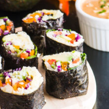 Several slices of vegan sushi with the dark nori on the outside sits by a bowl of spicy sweet peanut butter sauce.