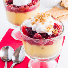 Two serving dishes full of vegan cherry vanilla parfaits are topped with whipped cream. Spoons sit beside them and graham crackers and more whipped cream is behind them.